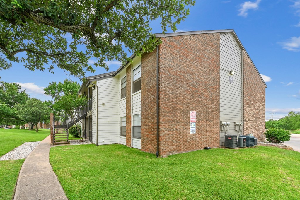 An apartment building with white wood and brick siding is surrounded by a lush grassy area at Estelle Creek North Apartments in Irving, TX