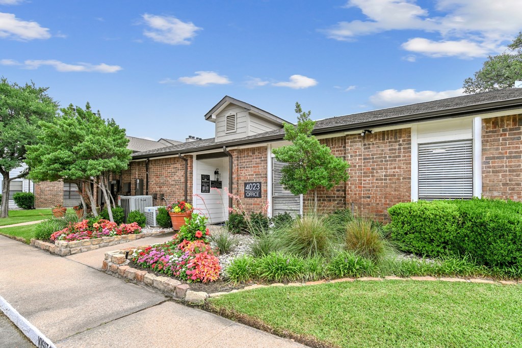 A leasing office surrounded by a well-kept lawn, lush trees, and blooming flowers at Estelle Creek North Apartments in Irving, TX