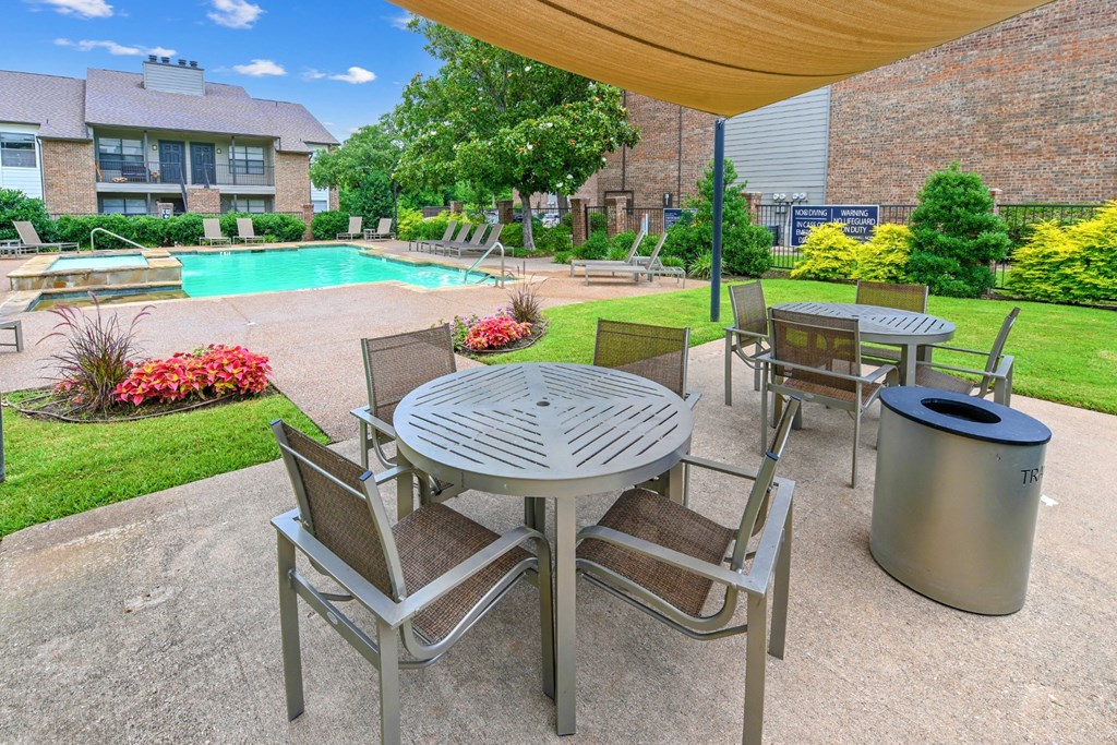 An outdoor dining patio with tables and chairs with a swimming pool in the background at Estelle Creek North Apartments in Irving, TX