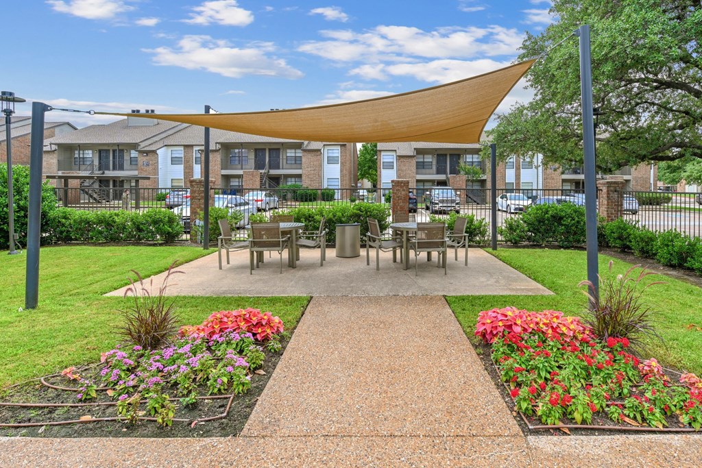 A shaded outdoor dining area with tables and chairs with a swimming pool in the background at Estelle Creek North Apartments in Irving, TX