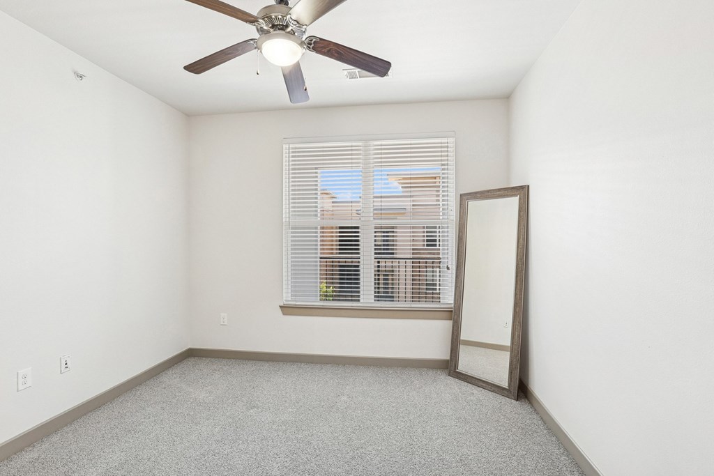 A bedroom with a ceiling fan, mirror, and a window with blinds at GreenVue Apartments in Richardson, TX