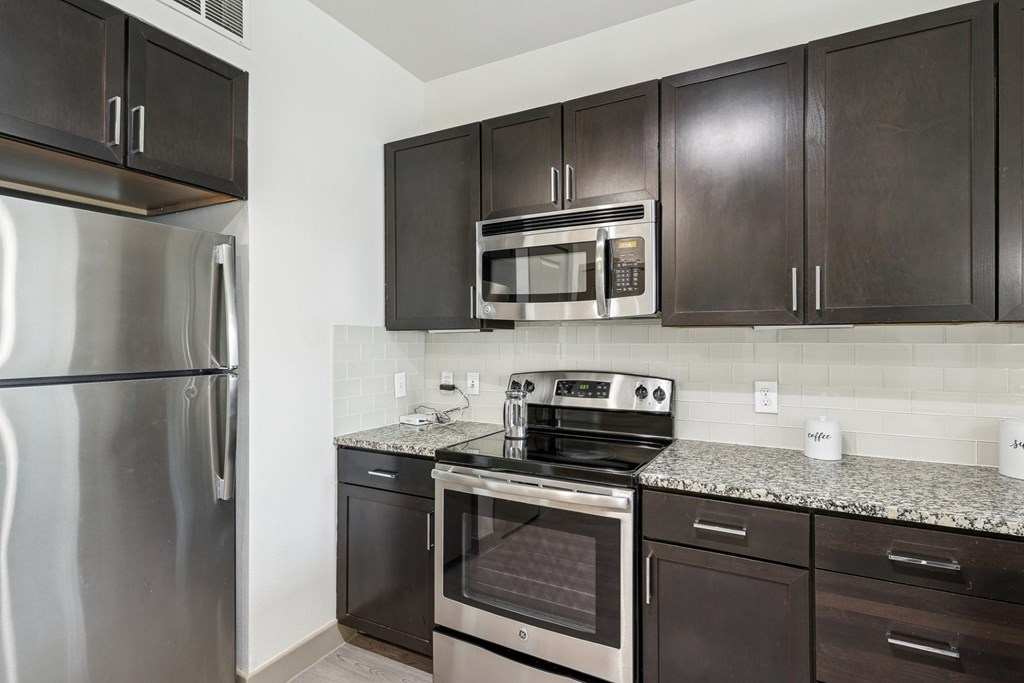 A kitchen with a stainless steel refrigerator, microwave, oven, and dark wood cabinets at GreenVue Apartments in Richardson, TX