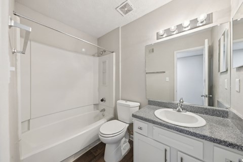 A bathroom with a white bathtub and shower, toilet, sink, and a mirror with lights at Gwinnett Square Apartments in Duluth, GA