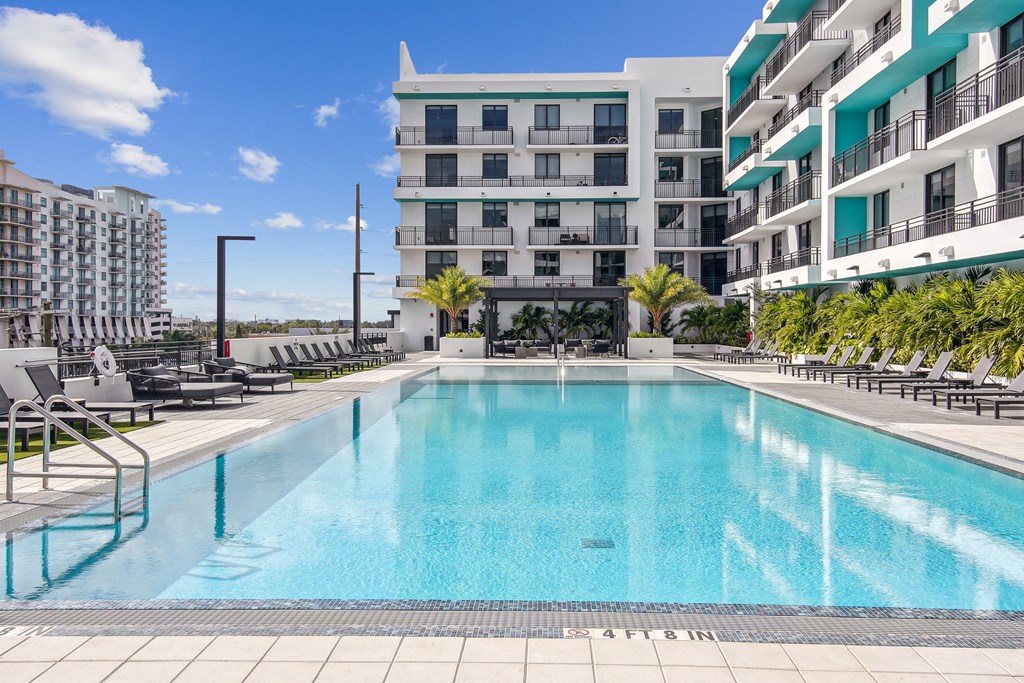 Large swimming pool surrounded by greenery and lounge chairs with a pergola in the background at Hollywood Heights on the Boulevard Luxury Apartments in Hollywood, FL