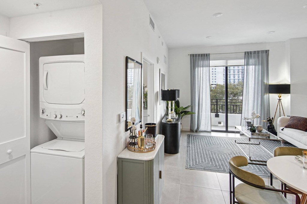 A modern living room with a window with open curtains, a dining table and chairs, and a washer and dryer  A modern kitchen with a dining table and chairs, fridge with a ice maker, stovetop, and microwave  Living room and modern kitchen in our 2-bedroom model with a dining table and chairs  Balcony view of Hollywood Boulevard at Hollywood Heights on the Boulevard Luxury Apartments in Hollywood, FL