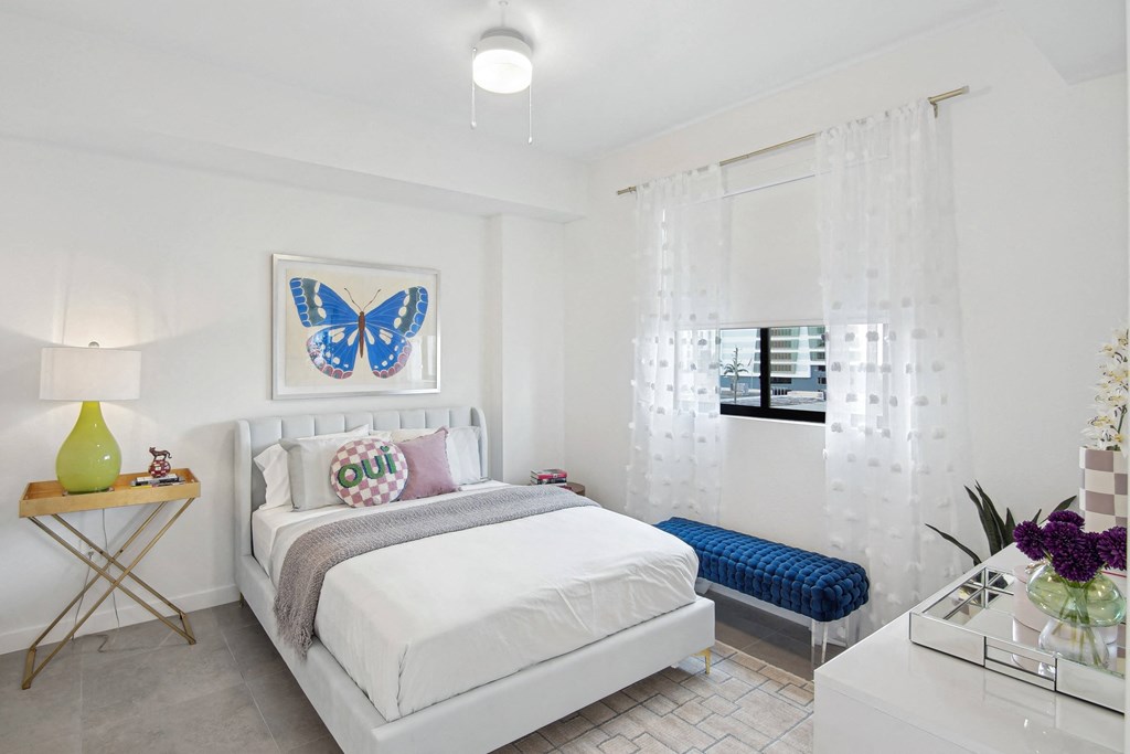 A bedroom with a bed, a blue bench, and a blue butterfly picture on the wall above the bed  Second bedroom with a bed, nightstand in our 2-bedroom model at Hollywood Heights on the Boulevard Luxury Apartments in Hollywood, FL