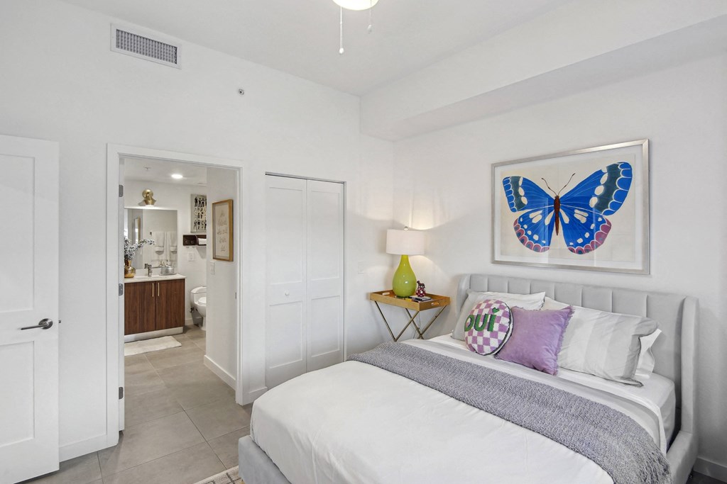 A bedroom with a bed, a blue bench, and a blue butterfly picture on the wall above the bed  Second bedroom with a bed, nightstand in our 2-bedroom model at Hollywood Heights on the Boulevard Luxury Apartments in Hollywood, FL