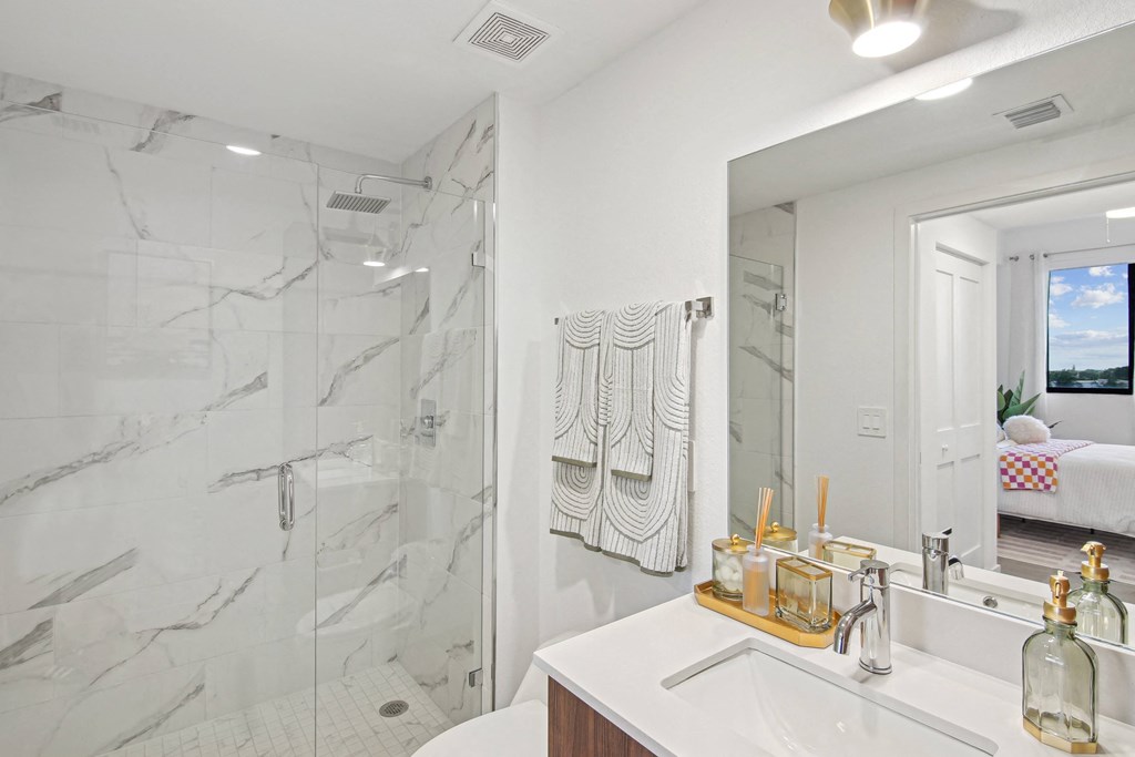 A bathroom with a marble shower and white countertop  A bedroom with a bed, a blue bench, and a blue butterfly picture on the wall above the bed  Second bedroom with a bed, nightstand in our 2-bedroom model at Hollywood Heights on the Boulevard Luxury Apartments in Hollywood, FL