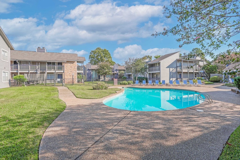 A swimming pool with a walkway and grassy area with apartment buildings in the background at Laurel Parc apartments in Shreveport, LA.