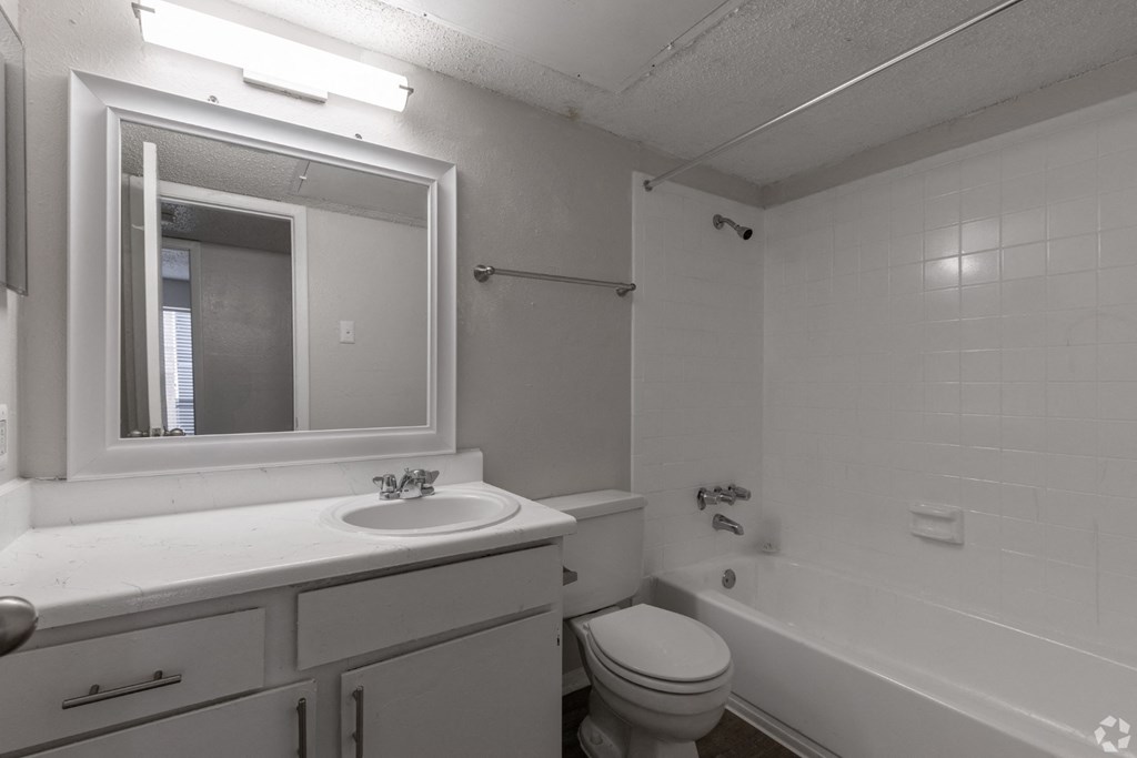 A white bathroom with a toilet, sink, mirror, and bathtub at Preston Oaks Apartments in Dallas, TX.