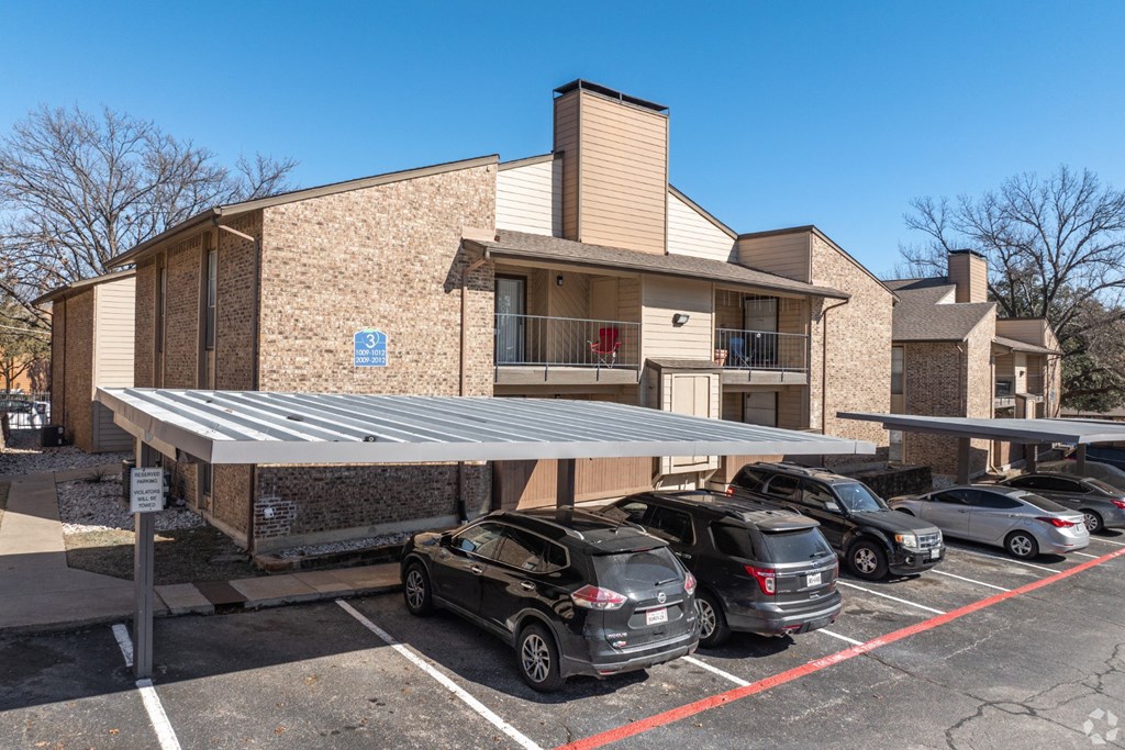A covered parking area with cars and an apartment building in the background at Preston Oaks Apartments in Dallas, TX.