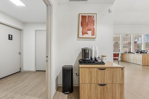 A room with a wooden floor, a white wall, a black trash can, a wooden cabinet with a black coffee maker on top, and a framed artwork on the wall.