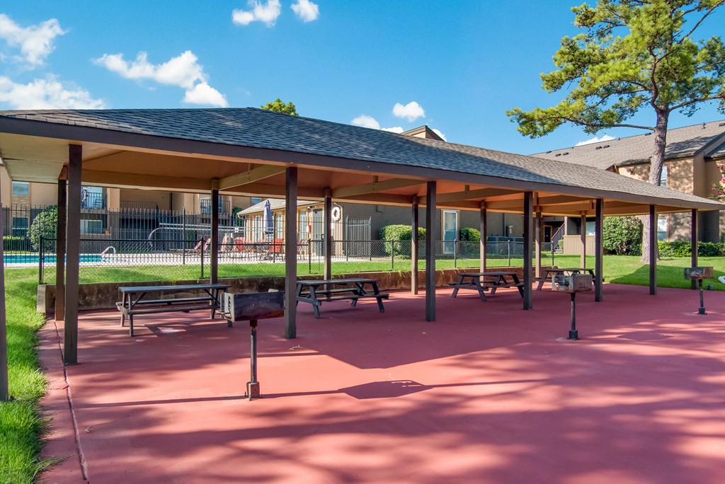 A covered picnic area with tables and benches and the pool in the background at Stone Canyon apartments in Shreveport, LA.