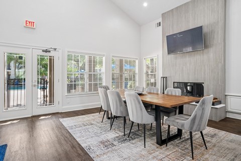 A clubhouse with a wooden table and chairs, a television on the wall, and a door leading outside at La Costa Apartments in Plano, TX