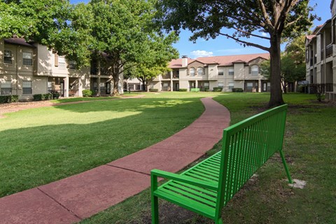 Walking path between apartment buildings with a green bench at La Costa in Plano, TX