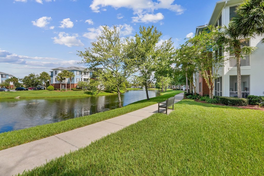 A walkway with a bench and a pond in front of apartment buildings at Wynnfield Lakes Apartments in Jacksonville, FL