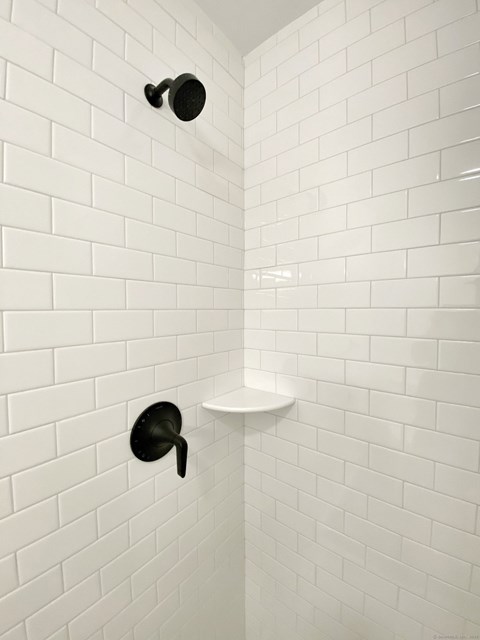 A white tiled shower with a black shower head and a white shelf.