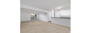the living room and kitchen of an apartment with white walls and carpet