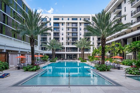 A large swimming pool surrounded by palm trees and lounge chairs in front of a multi-story building.