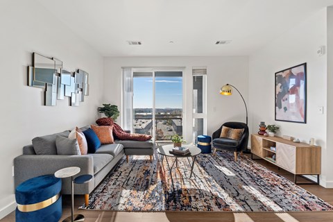 A living room with a grey couch, a blue ottoman, a wooden side table, and a large window with a view of the city.