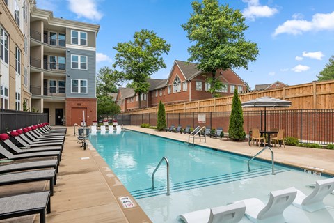 A pool with chairs around it and apartment buildings in the background.
