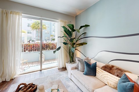 A living room with a couch, a plant, and a view of a balcony with chairs and a table.