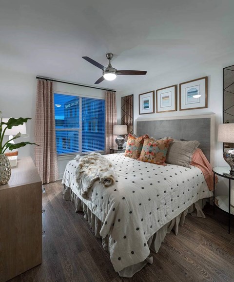 A bedroom with a bed covered in a white comforter with black dots, a ceiling fan, and a nightstand with a lamp.