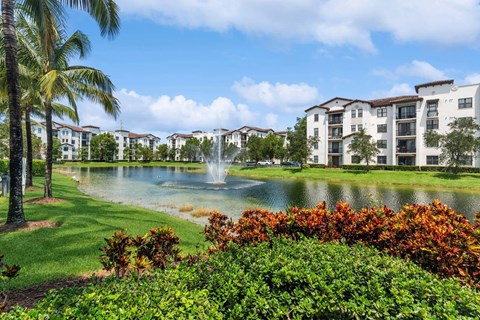 A beautiful view of a resort with a fountain in the middle of a lake surrounded by trees and buildings.