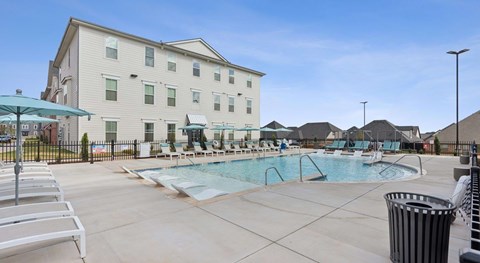 A large swimming pool is surrounded by lounge chairs and umbrellas in front of a white building.