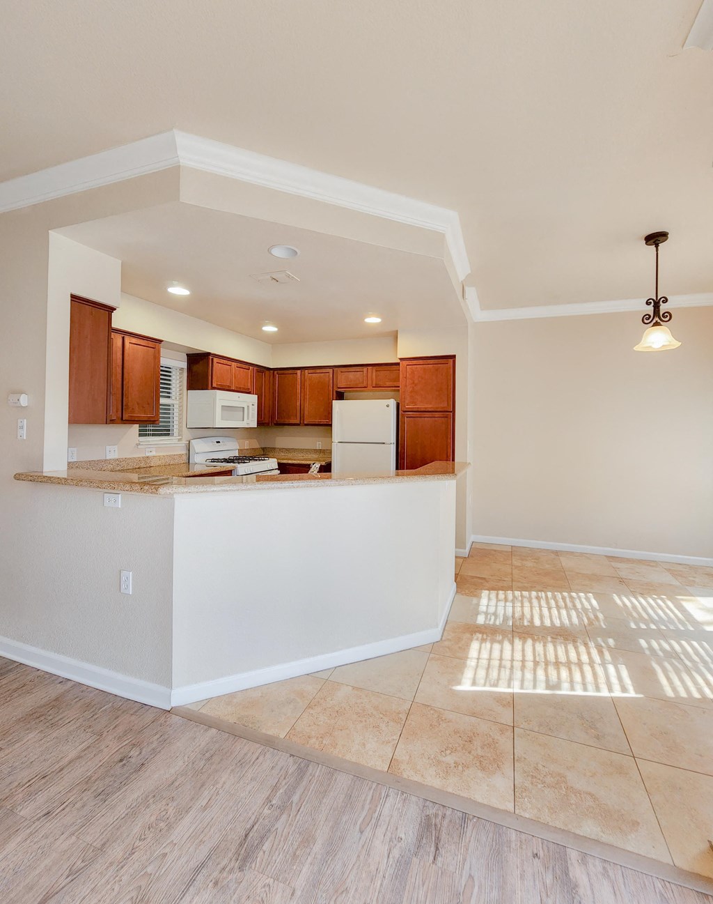 View from living area into kitchen and dining area. Brown cabinets, white appliances, tile flooring and wood look flooring, recessed lighting, wrap around kitchen counter.