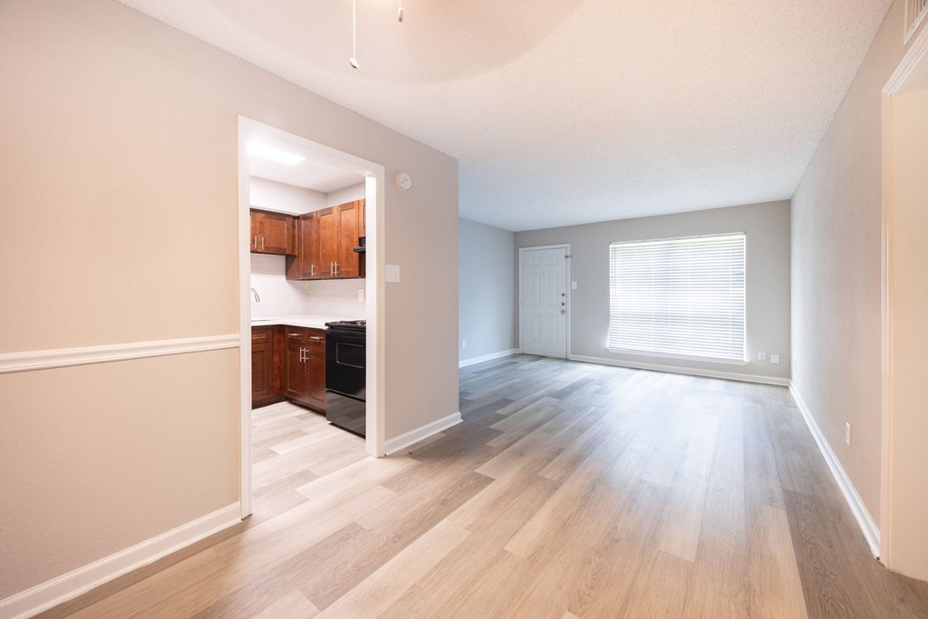 A room with a kitchen area in the background and a large window with blinds.