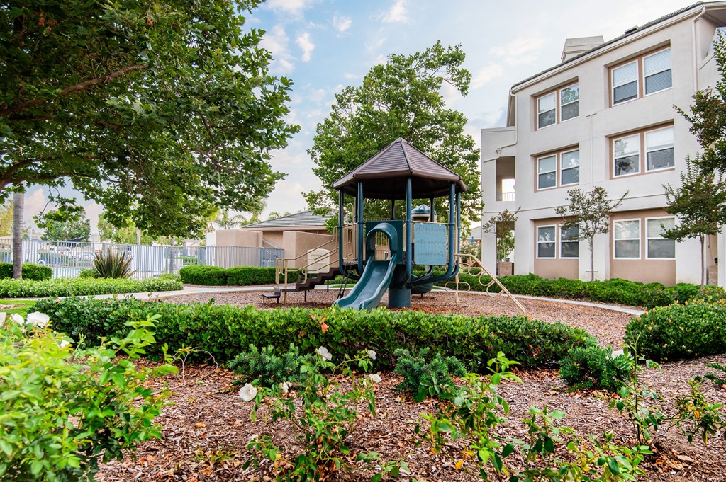 A playground with a green slide and a brown roof is surrounded by bushes and trees.