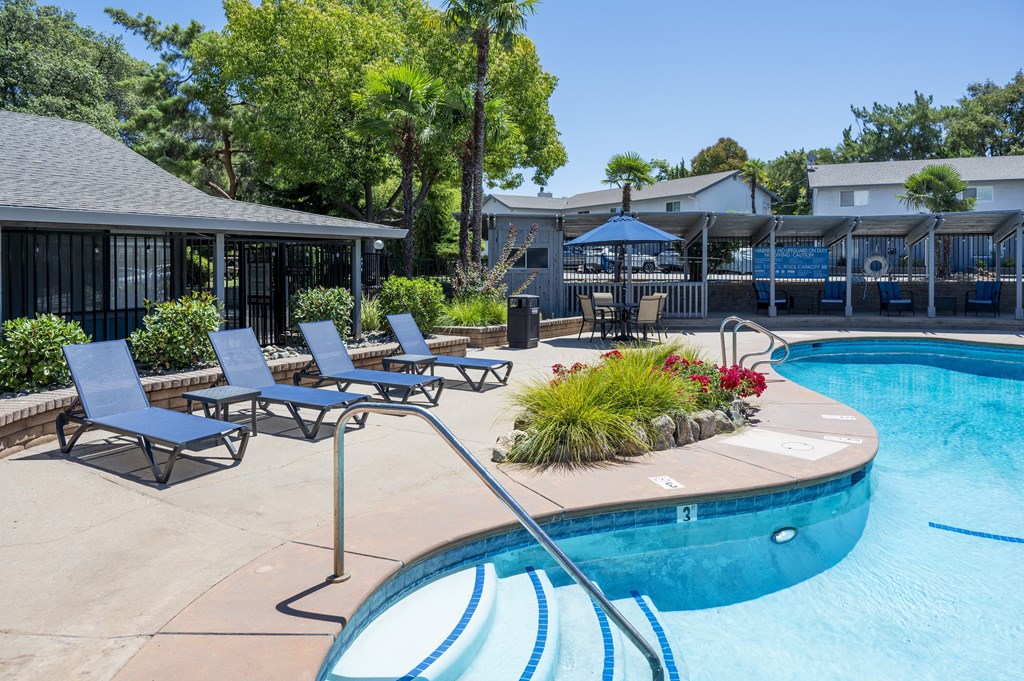 A pool with a blue tinted water surrounded by a concrete floor and a few chairs.