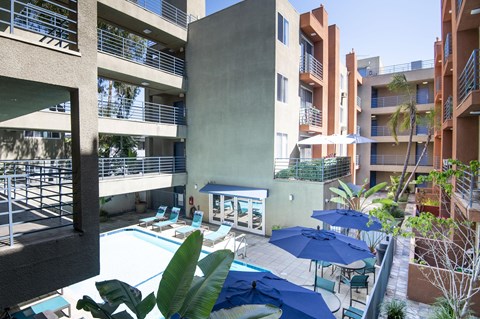 A pool area with blue umbrellas and chairs is surrounded by apartment buildings.