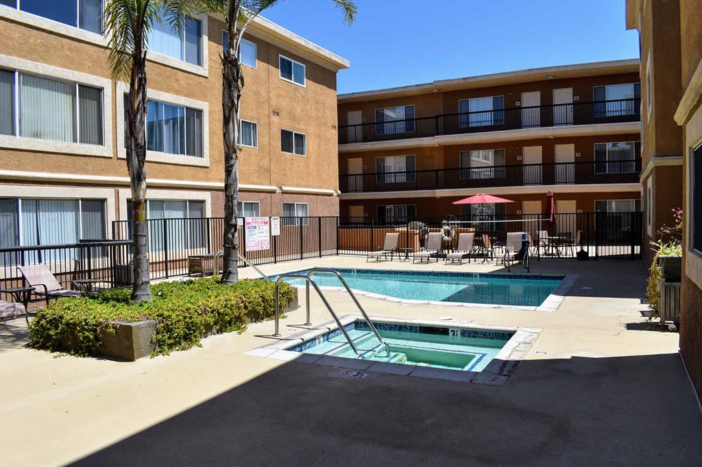 View of gated community pool and spa with lounge chairs and seating under an umbrella. Exterior of building in background