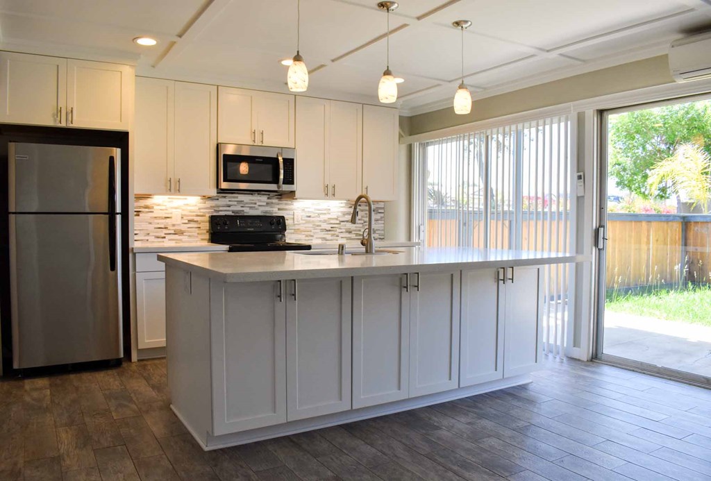 View of kitchen with island. Featuring wood look flooring, white countertops, white cabinets, stainless appliances, and tile backsplash.  Access into private patio/balcony through sliding glass door