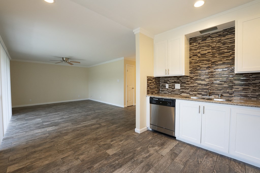 View from kitchen into living room, shows stone counters, tile backsplash, stainless appliances, white cabinets, wood look flooring and ceiling fan