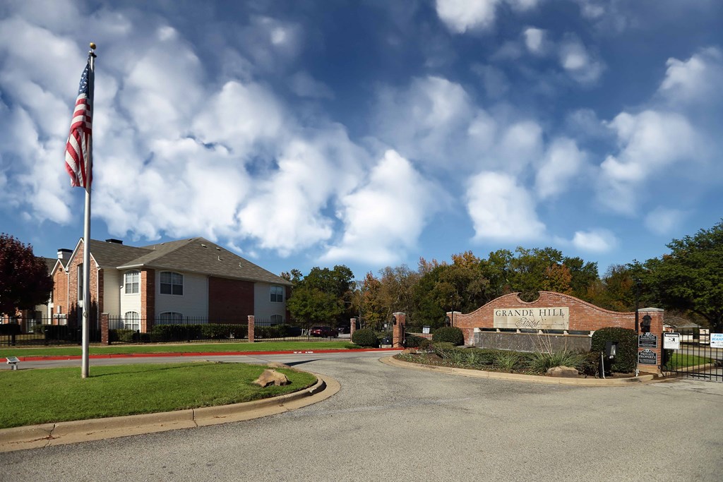 View of front entry gate with engraved Grande Hill Estates logo engraved in front entry water fall flower bed. Shows exterior building and American flag in back ground.