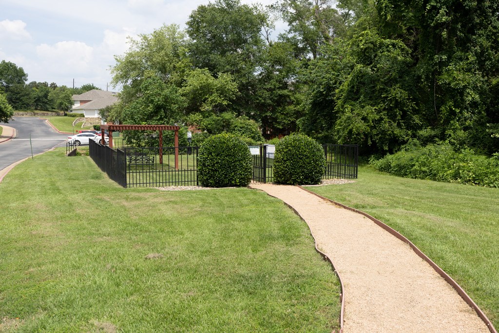 View of gated dog park with pergola and seating underneath and wooded area in background