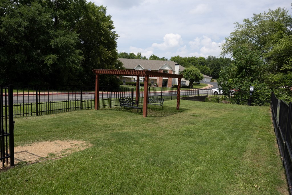 View from in gated dog park with pergola and seating area with wooded area and building in background