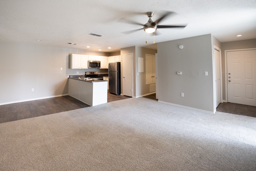 View of living area and open kitchen. Featuring carpet flooring in living area and wood look flooring in kitchen and dining room, with stainless appliances and dark counters.