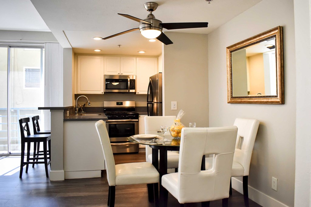 View of model kitchen and dining area with white cabinets, stainless appliances, dark counters, wood look flooring, bar on other side of counter, and dining table and chairs
