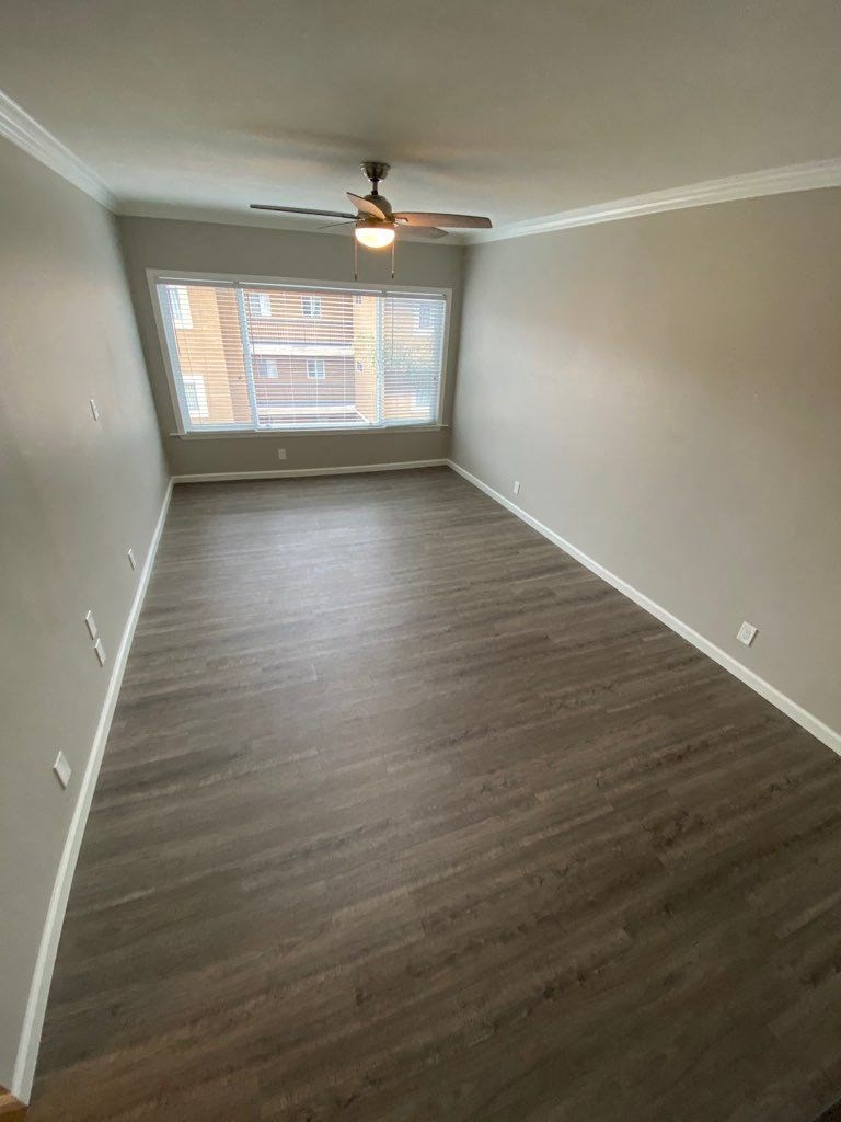 View of room with wood look flooring, ceiling fan, and well lit almost floor to ceiling window.
