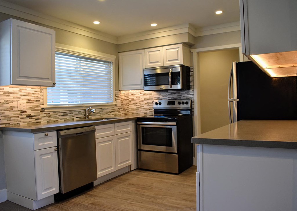 View of kitchen featuring stainless appliances, white cabinets, tile back splash, window above the sink and wood like flooring.