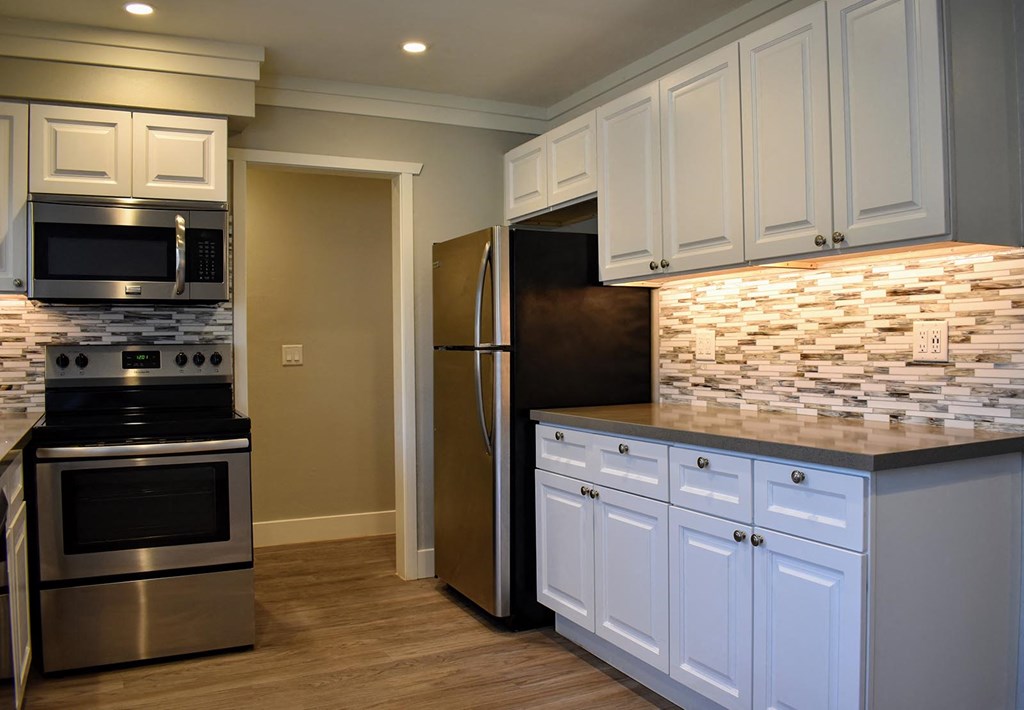 View of kitchen featuring stainless appliances, white cabinets, tile back splash and wood like flooring.