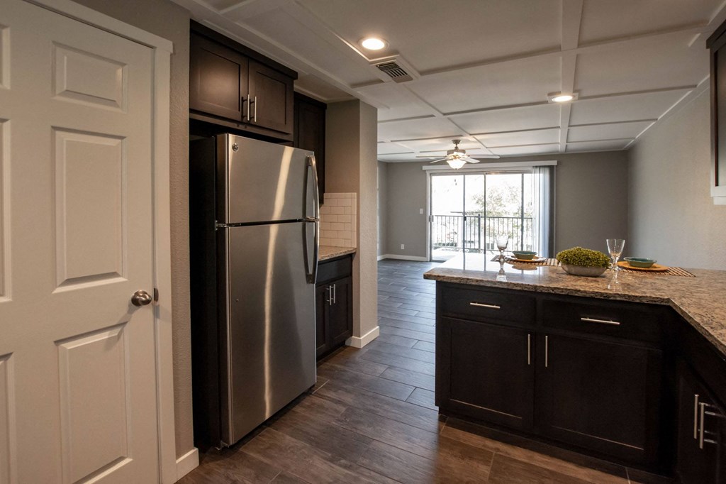 view from kitchen to living room wth wood look flooring, stainless appliances, tile backsplash, stone counters, ceiling fan, and private patio/balcony access via sliding glass door