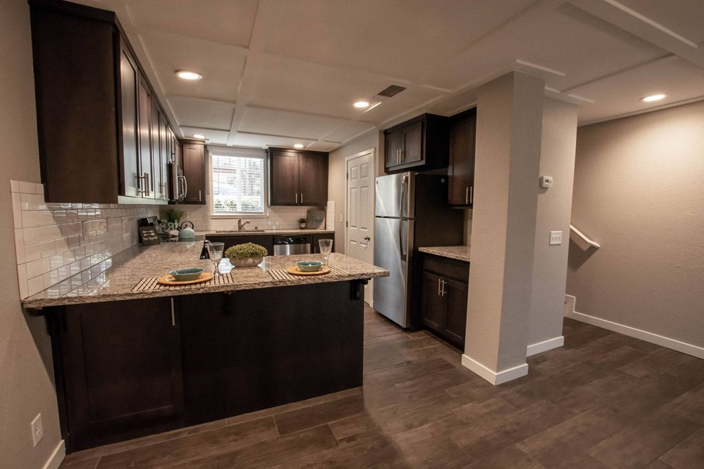 View from dining room into kitchen and partial view of stairwell to upstairs. Featuring stone counters, tile backsplash, brown cabinets, stainless appliances and wood look flooring