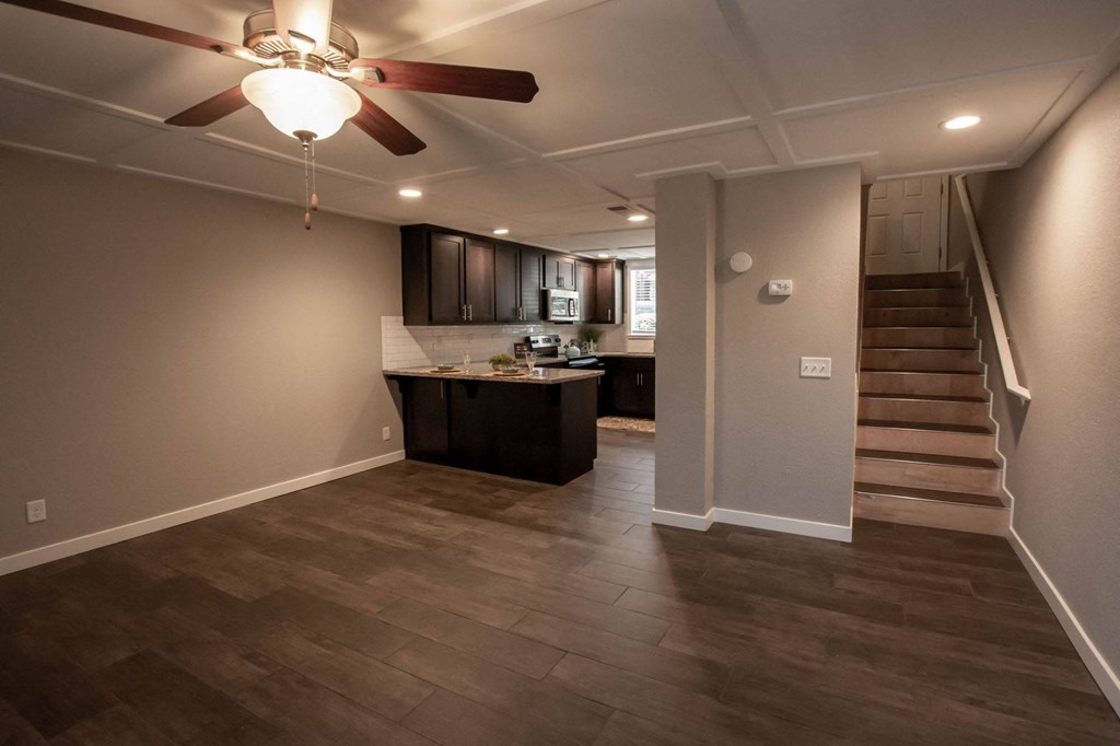 View from living and dining area into kitchen and up stairs in town homw, featuring wood look flooring, stone counters, tile backsplash, stainless appliances, brown cabinets, veiling fan and stairs leading upstairs