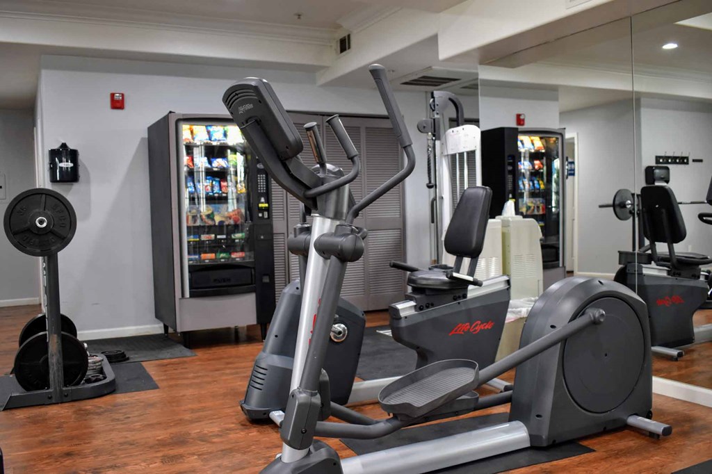 View of community fitness center with elliptical, stationary bike, squat rack, and vending machine