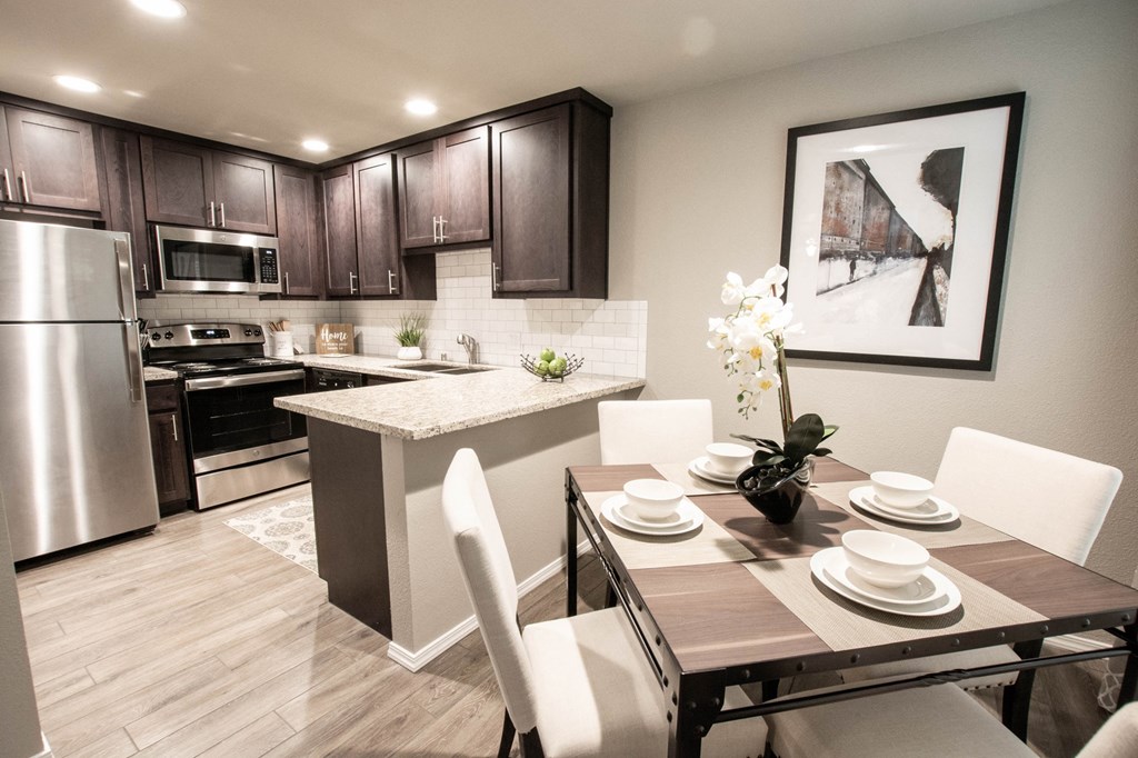 view from dining area into kitchen with wood look flooring, stone counters, granite counters, brown cabinets, stainless appliances, and tile backsplash